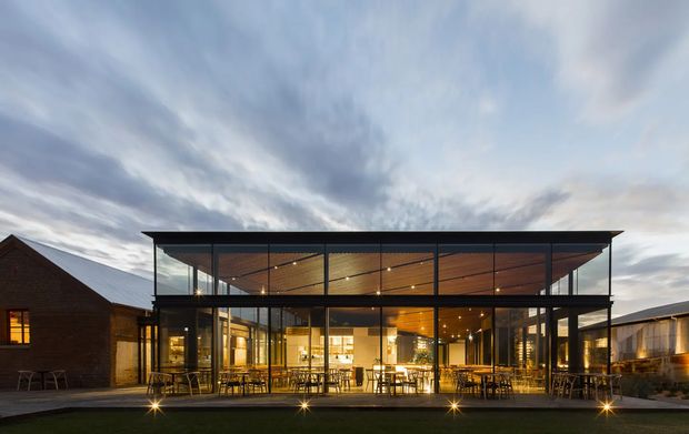 A dusk exterior photograph of a contemporary glass-fronted pavilion or extension, with a broad flat roof and dark structural framing. Warm timber-lined soffits and illuminated interiors are visible through extensive floor-to-ceiling glazing. Outdoor dining tables sit along the frontage, while older brick buildings flank the modern central volume.