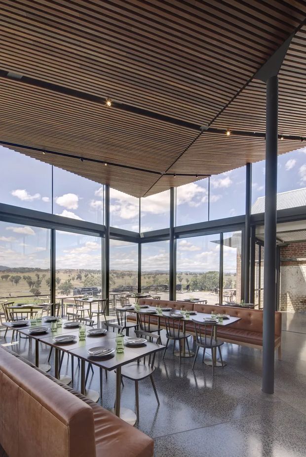 A contemporary restaurant or hospitality dining space with extensive floor-to-ceiling glazing overlooking an open rural landscape. The interior features rows of dark-framed windows, polished concrete flooring, long brown leather banquette seating, simple dark dining tables and chairs, and a dramatic suspended ceiling of closely spaced timber slats supported by dark steel columns and beams. The bright, airy room combines warm timber and leather with a cool industrial structural palette.