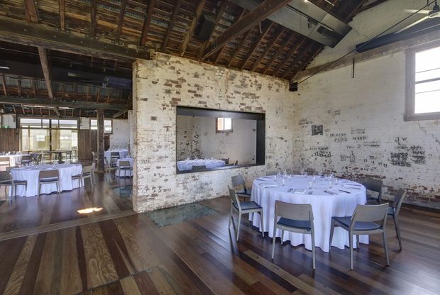 A rustic industrial event or dining space in a converted heritage-style building. The room features exposed timber roof framing and rafters, weathered white-painted brick walls, dark polished timber floors, large windows, and round tables dressed in white tablecloths with formal place settings. A large rectangular opening connects visually to an adjacent dining area, while suspended services and exposed structure reinforce the warehouse-conversion character.