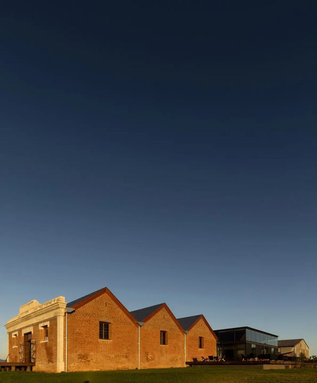 Exterior view of a long, low brick building complex under a clear deep-blue sky. The historic-looking red brick gabled wings feature repeated small windows and a prominent classical stone entrance surround, while a darker contemporary glazed extension connects at the rear. The building sits on an open lawn and is photographed in warm late-day light.