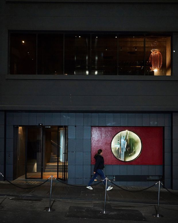 Night-time street-level view of a dark, contemporary building facade. The lower level has charcoal square cladding panels, a glazed entrance with visible stairs, and a large red feature panel containing a circular mirror or window. A long upper-level window reveals a warmly lit interior with a sculptural pendant light. A pedestrian and temporary rope barriers appear in the foreground.