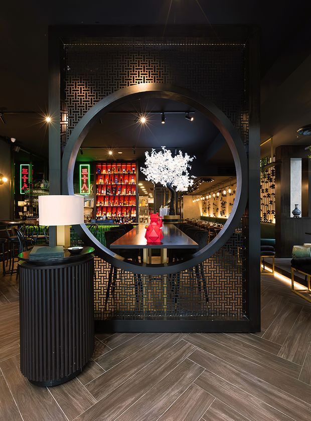 A dramatic, dimly lit restaurant or bar interior viewed through a large circular opening in a black decorative geometric screen. The space features dark finishes, patterned timber-look flooring in a herringbone layout, a backlit liquor display, illuminated wall shelving, dining tables, pendant and spot lighting, and a striking white blossom arrangement with red decorative objects on a central table. The overall atmosphere is refined, high-contrast, and luxurious.