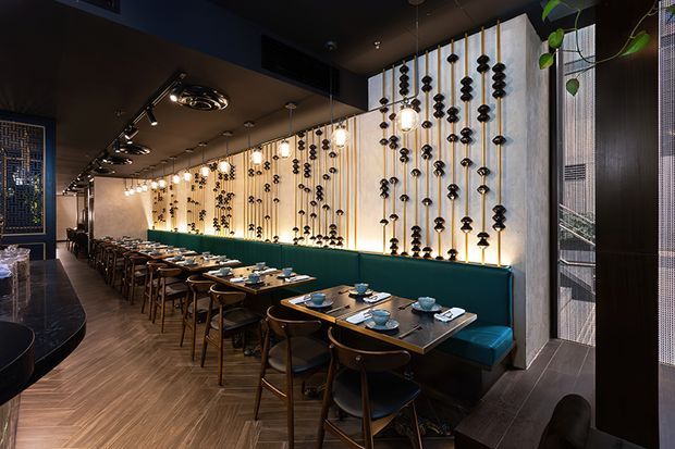 A long, sophisticated restaurant dining room with a continuous teal upholstered banquette, dark wood tables, and paired wooden dining chairs. A dramatic illuminated feature wall runs along the seating, decorated with vertical strands of black sculptural elements and pendant bulbs. The space has dark ceilings with track lighting, warm timber herringbone flooring, dark wall finishes, and a narrow reflective or glazed partition at the right. The overall atmosphere is intimate, moody, and refined.
