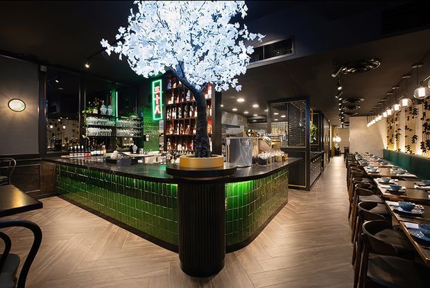 A sophisticated restaurant and bar interior viewed from the main dining area. A central island bar is clad in glossy deep-green rectangular tiles with dark ribbed detailing, surrounded by timber-look herringbone flooring. The bar features illuminated bottle displays, glassware, and a striking illuminated white blossom tree. To the right, a long dining banquette and rows of tables sit beneath pendant lights and decorative wall-mounted wine storage. The atmosphere is dark, atmospheric, and refined with dramatic accent lighting.