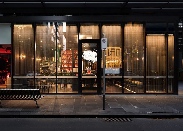 Street-facing glazed facade of a warmly lit hospitality venue at night, with dark metal framing, vertical screened blinds, bar shelving, tables and seating visible inside. The image is viewed from the footpath beneath a covered urban frontage.