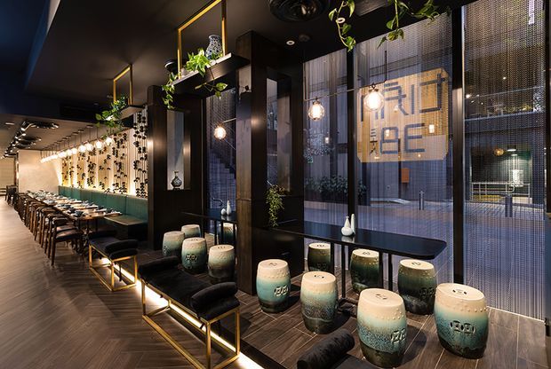 A stylish contemporary commercial dining and lounge interior, viewed along a long restaurant seating area. The space features dark timber or wood-look flooring, black ceilings and wall panels, floor-to-ceiling window screens, pendant globe lights, integrated shelving with plants, and a row of small upholstered stools and low seating. A long communal dining table with many chairs extends into the background, while decorative ceramic stools and dark lounge benches occupy the foreground. The atmosphere is warm, moody, refined, and urban.