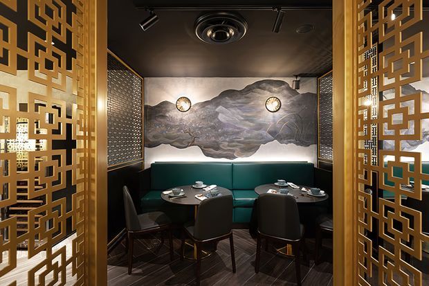 A dark, luxurious restaurant booth enclosed by ornate gold geometric screens. The space features a deep teal banquette, round black dining tables, upholstered dining chairs, dark timber flooring, black ceilings with track and recessed lighting, patterned wall panels, and a dramatic monochromatic mountain landscape mural with integrated wall lights.