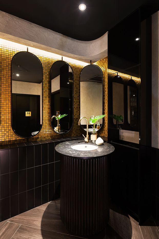 A dramatic, compact bathroom with a central round vanity featuring a white vessel basin, brushed gold tapware, and a dark fluted base. The walls combine glossy black lower tiles with small amber-gold mosaic tiles above, while tall arched mirrors and black cabinetry create a rich, reflective backdrop. A curved textured plaster ceiling and warm concealed lighting add sculptural character.