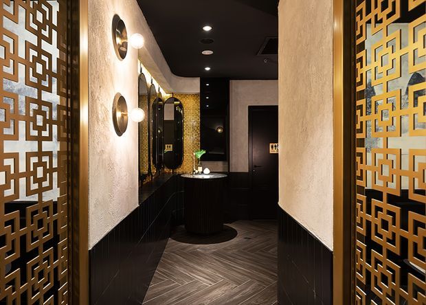 A dramatic, richly detailed interior corridor or bathroom approach featuring black lower wall panelling, textured cream plaster walls, a black ceiling with recessed downlights, and dark herringbone flooring. Decorative gold geometric screens frame both sides of the view. At the rear is a compact vanity area with a dark cylindrical basin unit, round mirrors, globe wall sconces, and gold-toned mosaic tile detailing. The overall aesthetic is dark, luxurious, and Art Deco-inspired.