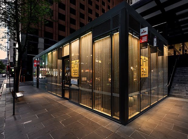 A compact contemporary commercial pavilion or storefront set within an urban streetscape. The structure has a dark metal frame, extensive floor-to-ceiling glazing, translucent golden mesh screening, warm concealed lighting, and large-format grey paving. Tall office buildings, street trees, signage, and an adjacent stairway surround the space.