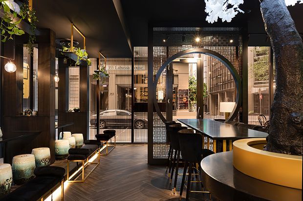 A sophisticated commercial hospitality interior, likely a restaurant or bar, viewed from a lounge seating area toward a long communal counter and service zone. The space features dark timber-toned finishes, black ceilings and walls, patterned metal or screen partitions, warm pendant lighting, floor-to-ceiling glazing, and a large circular portal framing the rear area. A substantial indoor tree sits in a curved mustard-yellow planter, adding an organic focal point. The atmosphere is moody, refined, and contemporary with strong geometric detailing.