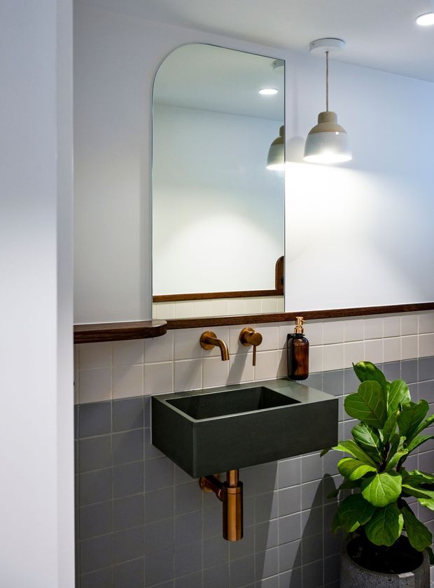 A compact, contemporary bathroom with a wall-mounted matte black rectangular basin, exposed copper-toned plumbing and taps, and a dark grey tiled backsplash with a horizontal white tile band. A large rounded-corner mirror, warm pendant light, timber-toned shelf trim, amber soap dispenser, and potted fiddle-leaf fig complete the space.