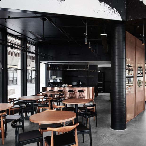 A dark, industrial-style commercial café or bar interior with exposed black ceiling services, large factory-style windows, concrete flooring, black metal-framed glazing, and a prominent cylindrical black structural column. The space contains numerous timber-top tables with black chairs, a black service counter with copper-toned fluted detailing, pendant lighting, and a timber-toned partition with circular display openings.