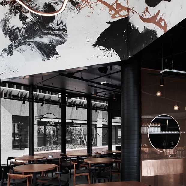 A stylish contemporary café or hospitality interior with dark industrial-inspired detailing, black-framed glazing, exposed ceiling services, pendant lighting, round timber tables, and black chairs. A large monochrome abstract mural spans the upper wall and ceiling, while a circular serving or display opening and metallic counter cabinetry feature on the right. The space opens visually toward an outdoor courtyard through extensive glazing.