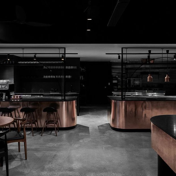 A dark, atmospheric commercial bar or hospitality interior with two copper-clad service counters, black stone benchtops, black metal-framed shelving and glass screens, pendant lights, bar stools, and tables. The polished industrial fit-out is set against a grey concrete floor and predominantly black walls and ceiling.