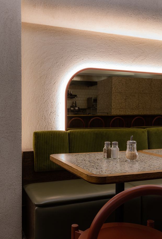 A warmly lit retro-inspired restaurant dining area with a built-in olive-green ribbed banquette, dark wood trim, rounded burgundy-toned chairs, and a rectangular terrazzo tabletop. An arched pass-through with integrated lighting reveals a tiled kitchen or service area behind it. The textured plaster walls and curved detailing create an intimate, refined atmosphere.