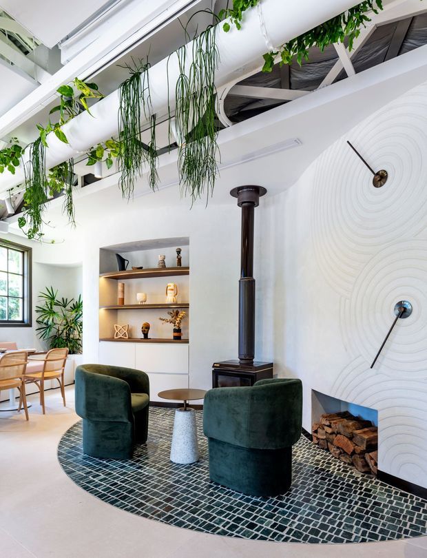 Bright contemporary living room with a sculptural white textured fireplace wall, black wood-burning stove, built-in display shelving, and two deep green velvet lounge chairs arranged on a curved mosaic tile rug. Exposed white ceiling structure and hanging greenery create an airy indoor garden feel, with a small dining area visible near the window.