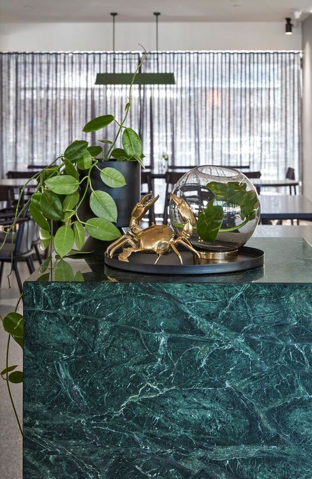 Close-up interior detail of a dark green veined stone-clad island or console with a polished matching benchtop. A black tray holds a trailing potted plant, a glass vessel, and a sculptural gold crab ornament. Sheer vertical blinds, dining furniture, and pendant lighting are visible in the background, creating a refined contemporary setting.