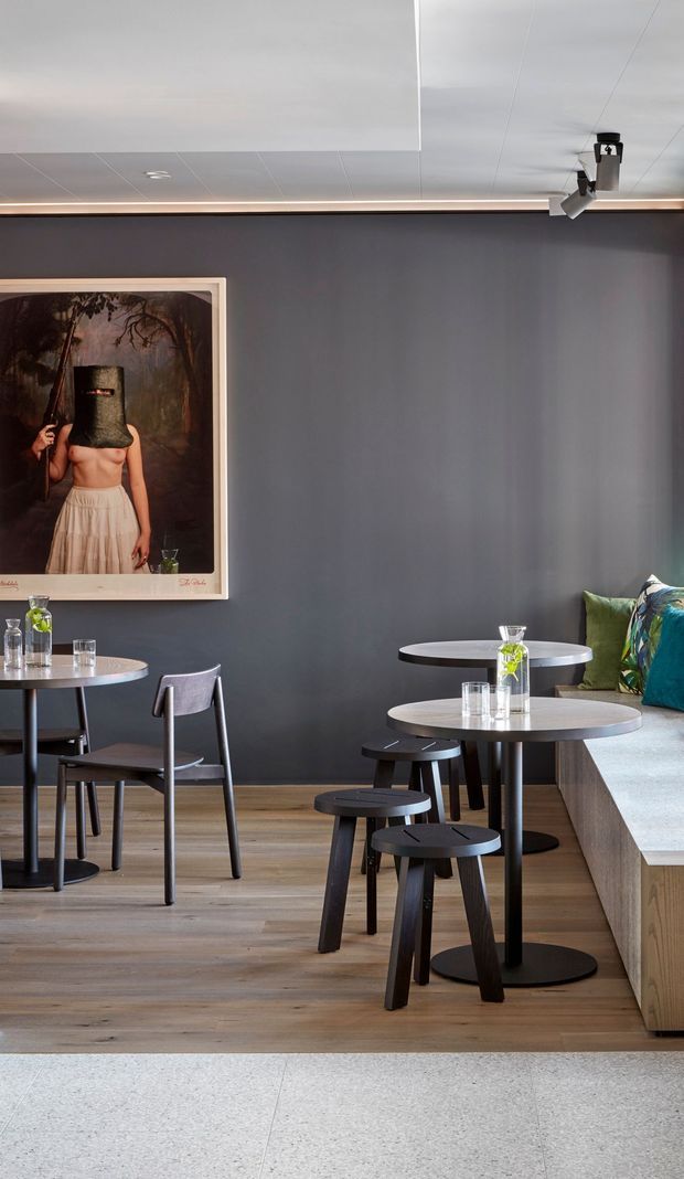 A refined café or hospitality interior with dark charcoal-grey walls, warm timber flooring, and a built-in upholstered bench along the right wall. Small round café tables, black stools, and simple grey chairs create a compact dining area. Track lighting and concealed linear lighting run along the ceiling, while a large framed artwork provides a visual accent.