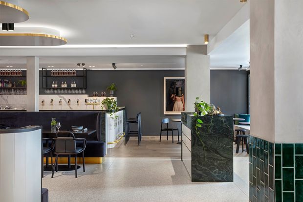 Contemporary hospitality interior resembling a stylish bar, café, or lounge. The space features a dark back bar with open bottle shelving, a compact service counter, black dining chairs and tables, pale timber flooring, terrazzo-like flooring in the foreground, and a dramatic dark green stone-clad island or reception counter. White walls and ceiling are complemented by integrated linear lighting, sculptural circular pendant lights with brass detailing, framed artwork, indoor plants, and glossy dark green tiled wall cladding.