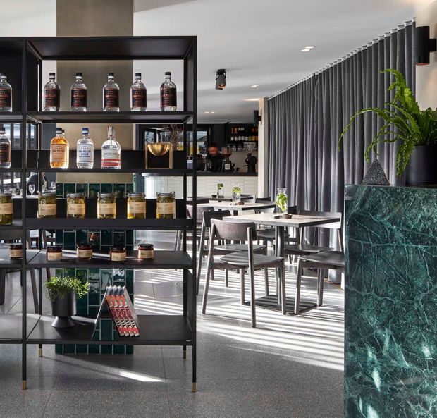 A contemporary bar or hospitality interior with a black metal display shelving unit stocked with liquor bottles and glassware in the foreground. Behind it are dining tables and upholstered chairs, a service counter, dark vertical drapery or fins, recessed lighting, and a striking green marble-clad bar or planter counter with greenery. Bright directional sunlight creates strong linear shadows across the terrazzo-like floor.