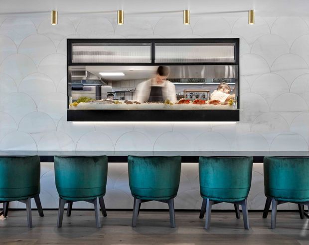 A refined restaurant or hospitality interior featuring a long counter with five teal upholstered chairs, a framed open kitchen service window, and chefs working behind the counter. The wall is clad in a pale scalloped patterned finish, complemented by dark trim, slim brass pendant lights, and timber-look flooring.