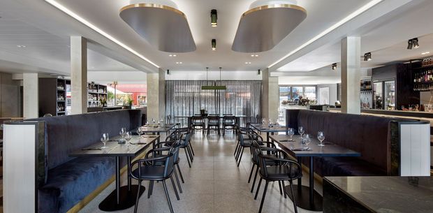 A contemporary restaurant dining room with symmetrical rows of black chairs and dark upholstered banquette seating arranged around timber tables. A central aisle leads to a glazed rear facade with vertical screening, while bar and bottle displays sit along both side walls. The space features a light tiled floor, white ceiling with linear LED lighting, sculptural metallic ceiling fixtures, and pale structural columns.