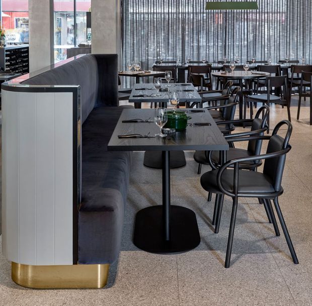 A contemporary restaurant dining area with rows of dark timber tables and black dining chairs, upholstered banquette seating, and a polished curved partition with a brass-toned base. Floor-to-ceiling windows are screened by vertical sheer blinds, while table settings and pendant lighting create a refined hospitality interior.