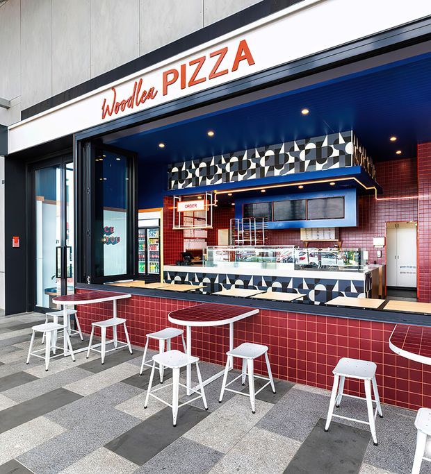 Exterior view of a contemporary takeaway pizza shop with an open service counter, bold red tiled facade, deep blue ceiling and trim, geometric black-and-white patterned wall tiles, illuminated menu and display areas, and outdoor bar-height tables with white stools.