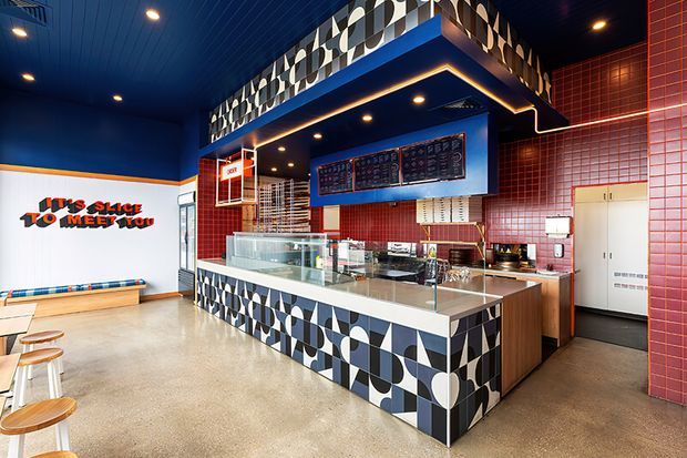 A brightly styled fast-food pizza shop interior with a prominent service counter, glass display case, menu boards, red tiled walls, deep blue ceiling and walls, and bold black-and-white geometric patterned cladding on the counter and upper fascia. Casual timber stools and bench seating line the left side, while playful wall lettering adds a branded feature.