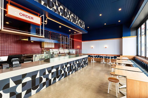 A brightly designed fast-casual restaurant interior with a prominent ordering counter, glass food display, open preparation area, wall-mounted menu boards, and rows of timber tables with stools along the window. The space features bold geometric black, white, and grey patterned tiles on the counter and upper wall, deep blue ceilings and walls, red tiled backsplash areas, warm timber seating, and polished concrete-look flooring. Large windows provide abundant daylight.