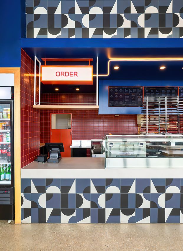 A brightly lit commercial food-service counter with a strong geometric mid-century-inspired fit-out. The storefront features bold blue, black, white and grey patterned wall panels, glossy red small-format tiled walls, a prominent illuminated ORDER sign, blue cabinetry or wall cladding with warm linear lighting, menu boards, refrigerated display cases, and stainless-steel food-service equipment. The image is photographed straight on from the customer area, with a drinks refrigerator visible at the left edge.