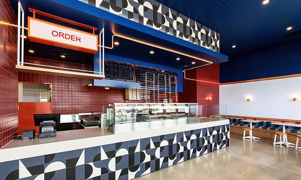Vibrant modern quick-service restaurant interior with a prominent ordering counter and glass food display, red tiled walls, deep blue ceiling and upper walls, and bold black, white, and grey geometric patterned cladding on the counter and upper fascia. Casual dining tables and stools occupy the right side of the space.