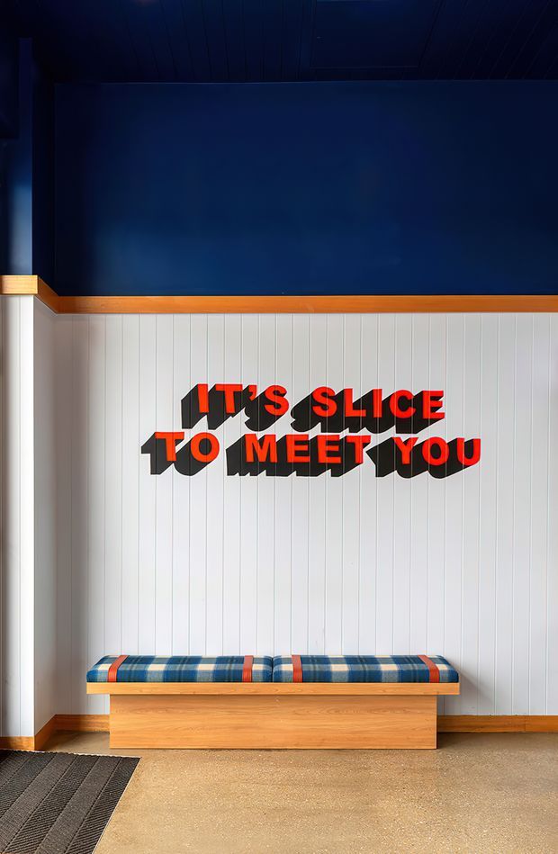 A compact commercial-style seating nook with a built-in timber bench against vertically grooved white wall panelling. The bench has blue plaid cushions with orange-red stripes and a bold dimensional red-and-black wall sign reading “IT’S SLICE TO MEET YOU.” A deep navy upper wall, warm timber trim, dark ribbed entrance mat, and polished concrete or terrazzo-like floor create a playful, branded, retro-inspired interior.