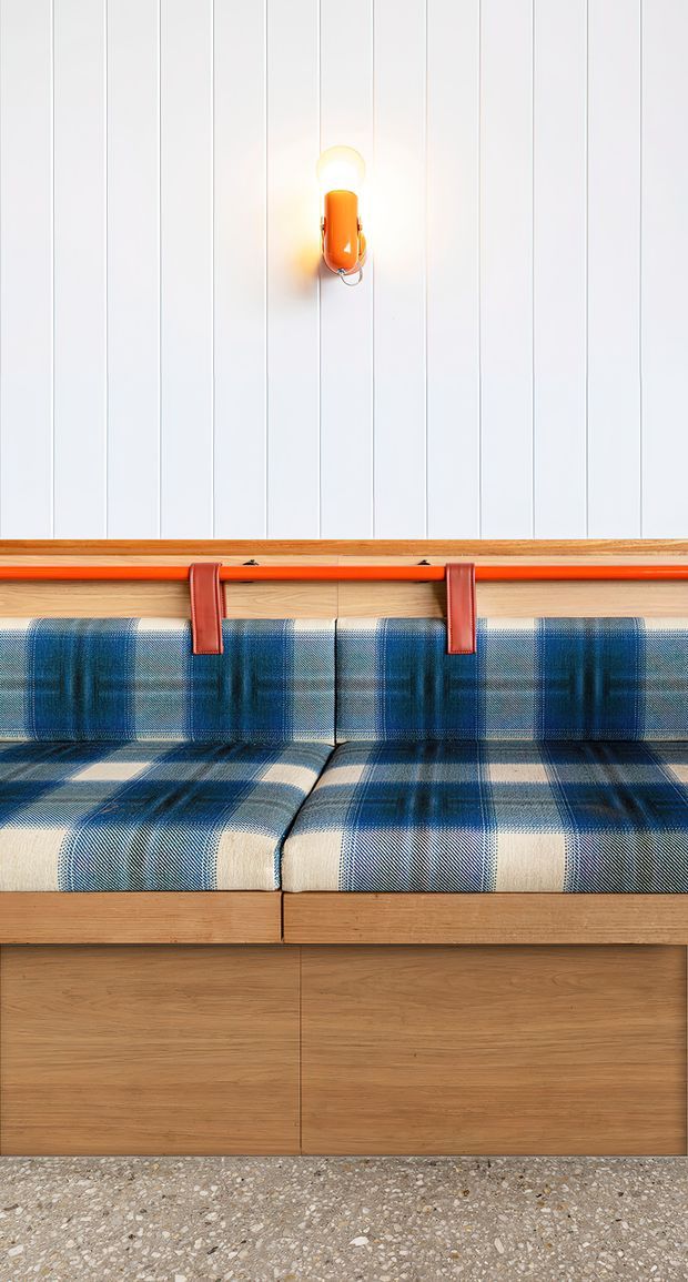A close-up of a built-in upholstered bench seat against a white vertically panelled wall. The bench has a blue and cream plaid fabric cushion and backrest, a warm timber base and ledge, and distinctive orange-red leather straps and trim. A small orange wall light is mounted above, while a terrazzo-style floor is visible below. The composition is brightly lit and strongly graphic, with a playful mid-century-inspired colour scheme.