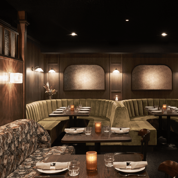 A dimly lit, intimate restaurant dining room with continuous curved olive-green upholstered banquettes, dark timber wall panelling, inset textured acoustic or decorative wall panels, dark ceilings, and multiple small dining tables set with crockery, glassware, folded napkins, and candlelit lanterns. Patterned upholstered armchairs and warm wall sconces contribute to a moody, refined hospitality interior.