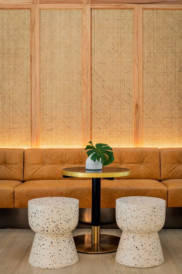 A warmly lit hospitality-style seating area with a long caramel-brown tufted leather banquette against a wall of natural woven cane or rattan panels framed in timber. A small round black-and-brass pedestal table sits in front, topped with a white planter and green tropical foliage. Two sculptural cream terrazzo stools with dark aggregate are positioned in the foreground over a pale timber floor.
