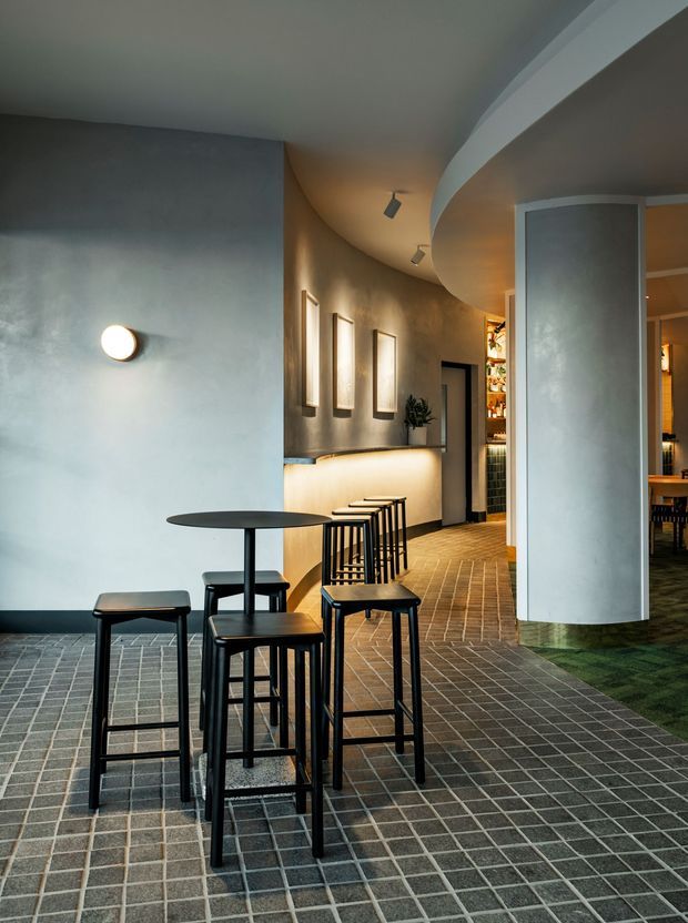 A moody contemporary commercial interior, likely a bar or restaurant, featuring a curved plastered wall and ceiling, dark gridded tile flooring, a slim black bar-height table with stools, softly illuminated wall-mounted joinery, framed artwork, and a partially visible dining area beyond.