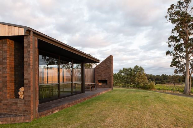 Exterior view of a low, contemporary brick house in a rural landscaped setting. The building features dark brick walls, timber-clad upper detailing, extensive floor-to-ceiling glazing, a broad low-pitched roof overhang, and a projecting brick outdoor fireplace or chimney element. A covered outdoor terrace with furniture is visible through the glazing and beneath the roof, opening onto a lawn and planted property boundary.