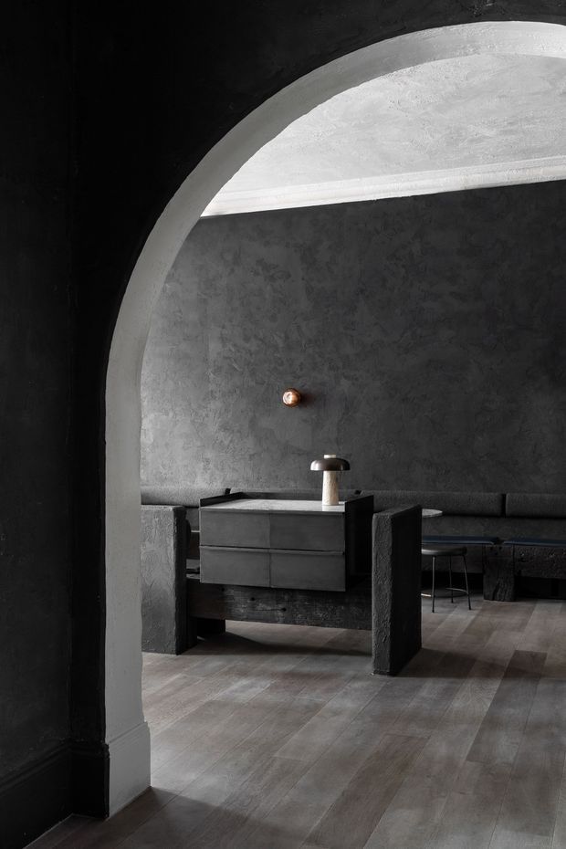 A moody monochrome interior viewed through a large rounded archway. The room contains dark textured plaster walls, a long built-in upholstered bench, a substantial dark timber console or dining table, and a compact black cabinet with a lamp. Grey-brown timber flooring and dramatic curved architectural detailing reinforce the sculptural, minimalist atmosphere.