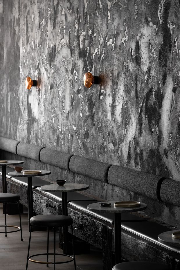 Moody monochrome commercial dining interior with a long upholstered banquette along a heavily textured dark grey feature wall. Small round black tables, black stools, dark flooring, and warm copper wall sconces create a refined, dramatic restaurant or bar setting.