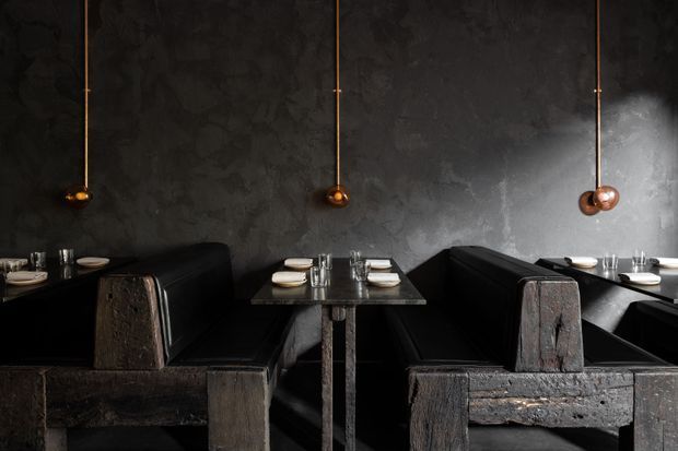 Dark, moody restaurant dining interior with rows of intimate booth seating, dark textured walls, black tables, and warm copper pendant lights. Tables are set with plates and glassware, creating a dramatic upscale hospitality atmosphere.