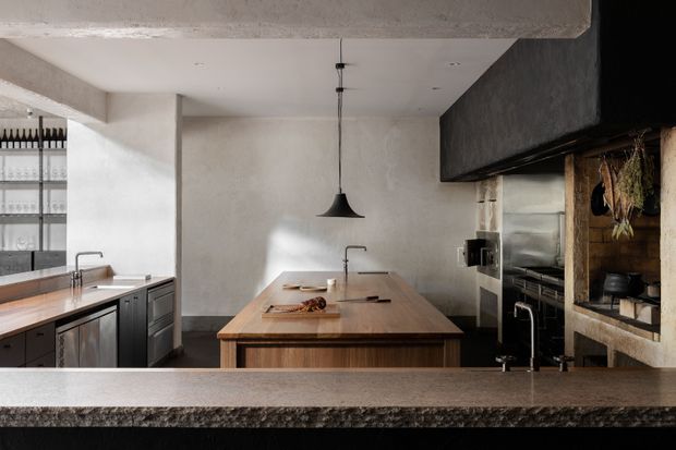 A sophisticated open-plan kitchen viewed across a dark stone or concrete foreground counter. The room features a large central timber island with an integrated sink and mixer tap, dark built-in cabinetry, a secondary timber worktop at left, exposed textured plaster or concrete walls, black pendant lighting, and a substantial open hearth or fireplace-like cooking area on the right. Industrial architectural detailing is balanced with warm timber surfaces and restrained contemporary styling.