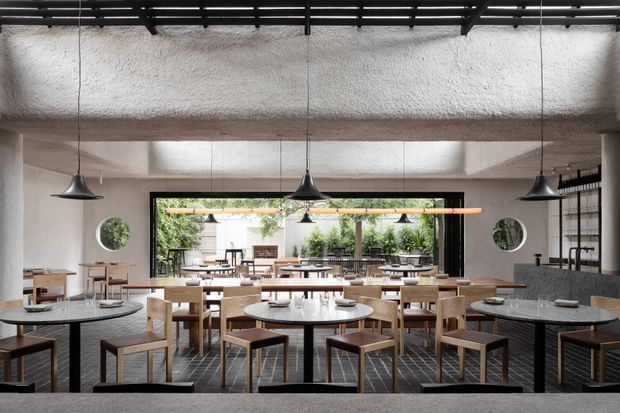A spacious contemporary restaurant or hospitality dining room with multiple round and rectangular tables, timber chairs, dark stone tabletops, and a tiled floor. Large black-framed glazed openings connect the dining area to planted outdoor greenery. The room features exposed concrete or plaster structural elements, black ceiling rails, and sculptural black pendant lights, creating a refined industrial-modern atmosphere.