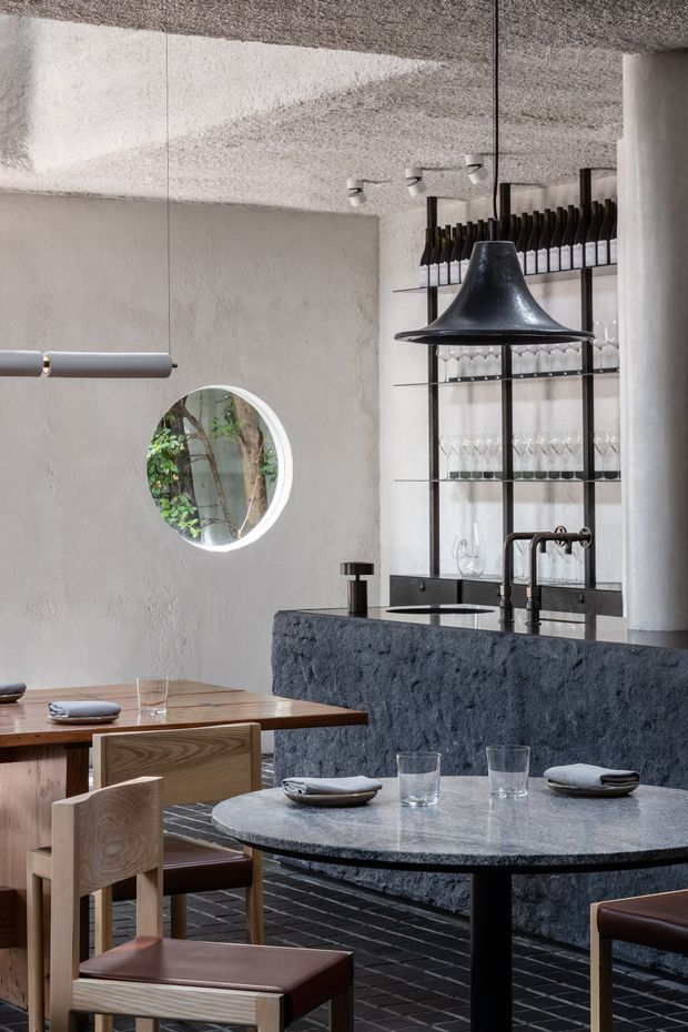 A sophisticated restaurant dining interior with a compact bar and wine display at the rear. The space features textured pale plaster walls and ceiling, a dark rough-textured bar front, open black metal shelving stocked with wine bottles and glassware, sculptural black pendant lighting, and a mix of round and rectangular timber dining tables with leather-upholstered chairs. A circular illuminated wall opening or mirror adds a distinctive architectural detail, while dark tiled flooring and charcoal surfaces create a refined, moody atmosphere.