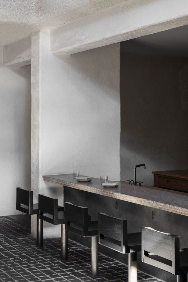 A dramatic, minimalist kitchen and dining counter with a long stone benchtop, dark timber cabinetry, black sculptural bar stools, and a black tiled floor. The space has white textured plaster walls, exposed structural beams, and a deep shadowed recess behind the sink, creating a stark, moody architectural interior.