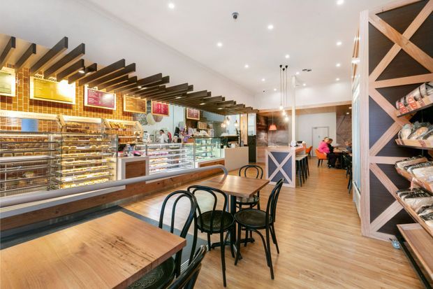 A bright bakery or café interior with a long display counter, illuminated menu boards, pastry shelving, timber dining tables, black bentwood-style chairs, and decorative exposed dark timber ceiling beams. The space combines warm timber flooring and furniture with white walls, yellow-brown tiled counter cladding, and geometric timber-framed retail shelving.