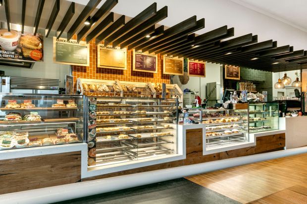 A bright contemporary bakery or café interior viewed from the customer area, with extensive glass pastry display counters, refrigerated food cases, timber-faced cabinetry, a tiled feature wall, menu boards, and a dramatic black exposed linear ceiling installation with integrated spotlights. Pendant lights and shelving continue toward the service counter at the rear.