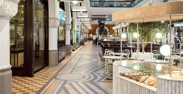 Interior view of an upscale covered retail and dining arcade, with patterned geometric mosaic flooring, ornate classical columns and decorative ceiling detailing. Boutique storefronts line the corridor, while a contemporary café kiosk with a pastry display, round globe lights, outdoor-style tables and chairs occupies the right side.