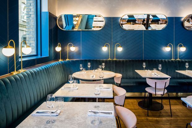 A refined restaurant dining interior with deep blue chevron wall panelling, teal upholstered banquette seating, pale stone dining tables, curved upholstered chairs, globe wall and floor lamps, and elongated mirrored wall fixtures. Timber flooring and warm lighting create a sophisticated, intimate atmosphere.